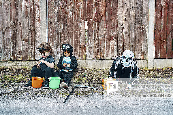 Drei für Halloween gekleidete Kinder  eines als Skelett  die mit ihren Eimern mit Süßigkeiten am Straßenrand sitzen.