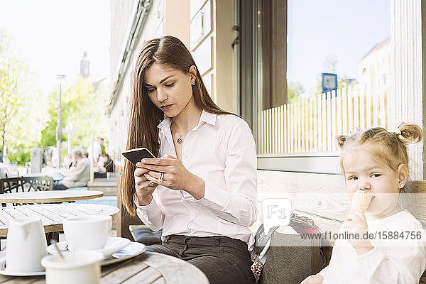 Eine Mutter und ihre Tochter sitzen vor einem Café  die Mutter am Handy  die Tochter isst ein Eis.