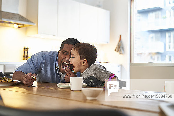 Ein Mann und ein Sohn an einem Küchentisch  beide mit weit geöffnetem Mund  essen ein Stück Schokoladenkuchen.