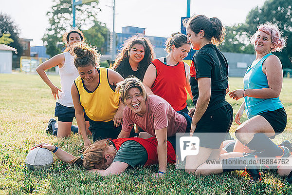 Eine Gruppe von Frauen kniend auf einem Rugbyfeld und lachend mit einer mit dem Gesicht nach unten auf dem Boden liegenden Frau.