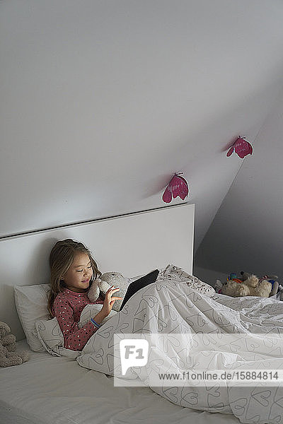 Home life  a school morning during lockdown. A girl lying in bed using a digital tablet.