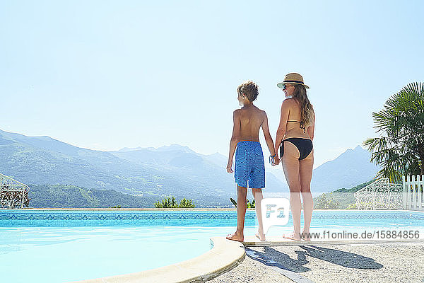 Eine Frau und ein Junge halten sich an den Händen und stehen neben einem Swimmingpool mit Blick auf die Berge in der Ferne.