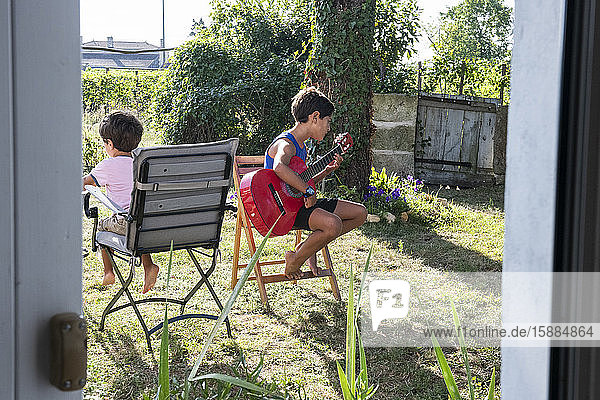 Zwei Jungen sitzen auf Stühlen im Garten  einer spielt Gitarre.