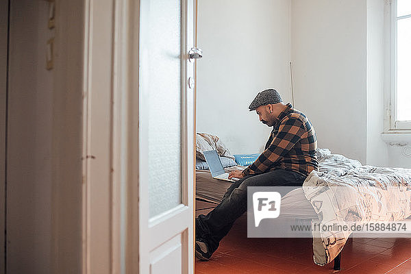 Mann mit dickem Schnurrbart und flacher Mütze saß während der Corona-Virus-Krise mit Laptop-Computer auf dem Bett.