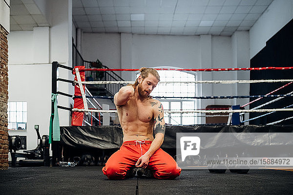 A bare chested man wearing red tracksuit trousers and kneeling on the floor in front of a boxing ring in a gym.