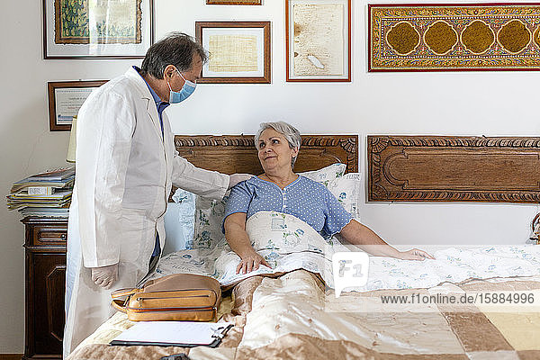 Ein Arzt im weißen Kittel und mit einer schützenden Gesichtsmaske bei einem Hausbesuch bei einer älteren Patientin.