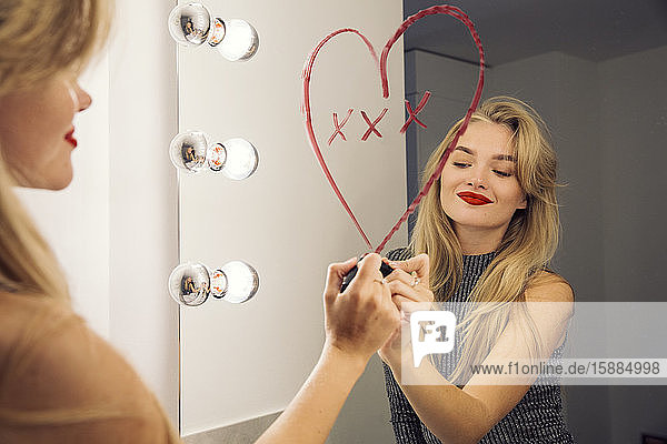 A woman looking in a bathroom mirror drawing a heart on the mirror with lipstick.