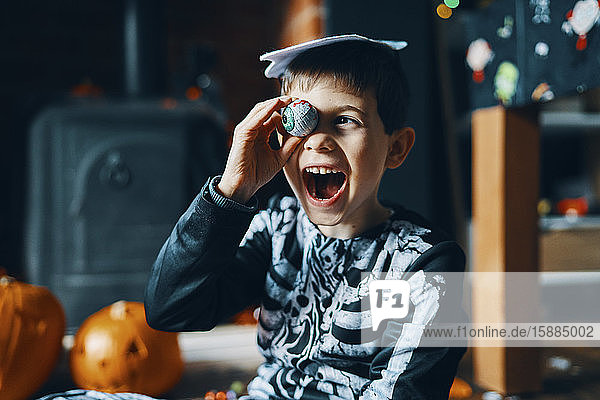 A boy dressed as a skeleton holding a chocolate wrapped as an eyeball to his eye.