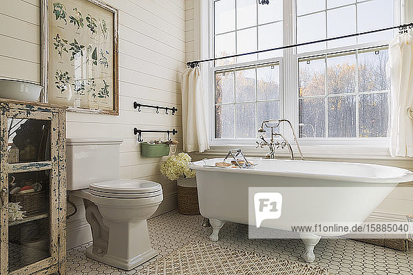 Bathroom with white roll top bath cream wood panelled wall and tiled floor sash windows with white curtains.