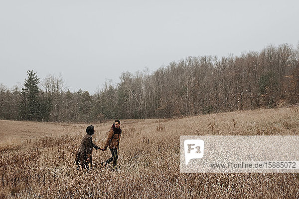 A couple holding hands and looking at each other walking through long grasses in a wintry landscape.