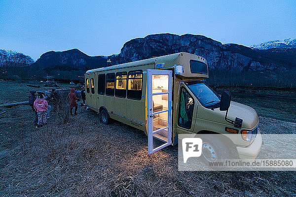 Campervan in a rocky landscape at dusk with the light on inside and the door open group of people standing towards the rear.