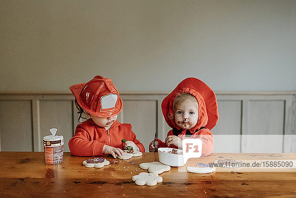 Two children dressed as firefighters at a table decorating cookies with chocolates and sprinkles.