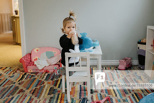 A child kneeling on a small white chair by a white table drinking from a bottle.