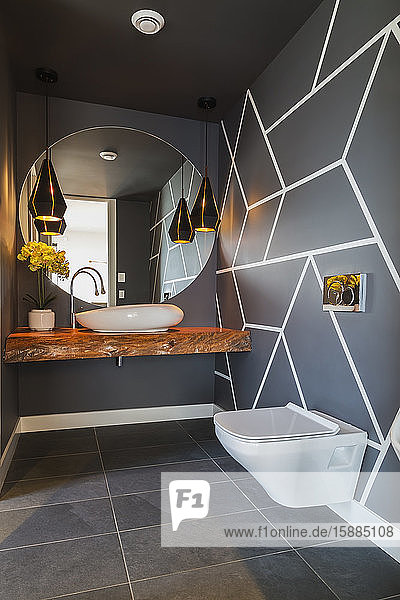 Interior view of modern bathroom with dark grey floor tiles and walls with geometric pattern round wall mirror over basin on wooden railway sleeper and wall mounted toilet.