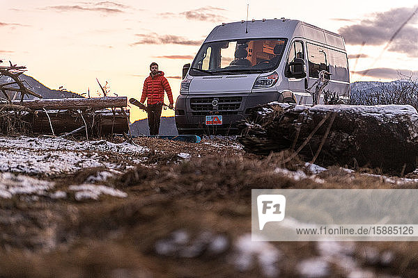 Man by a camper van carrying skis in a rocky wintry landscape.
