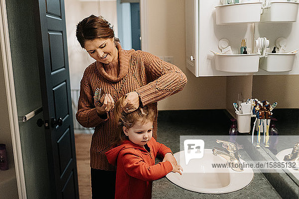 A woman brushing her daughters hair while standing in the bathroom.