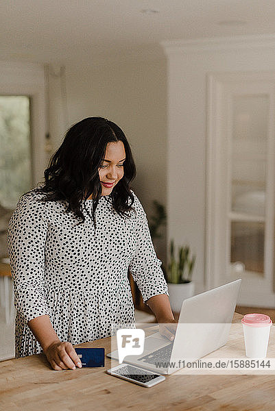 Woman with long dark hair standing at kitchen counter looking at laptop computer.