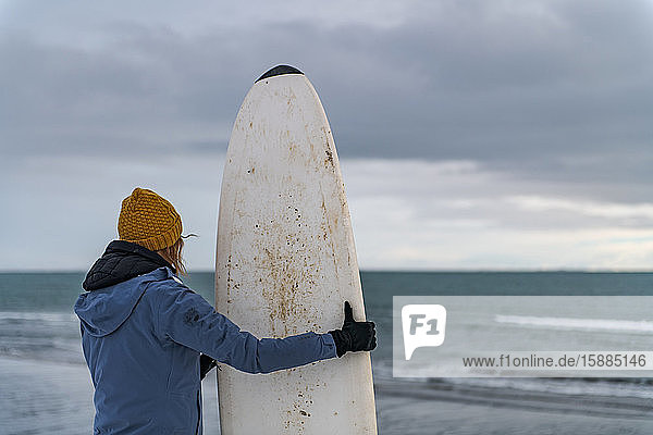 Rear view of a woman holding a surfboard standing on a snowy beach and looking out to sea.