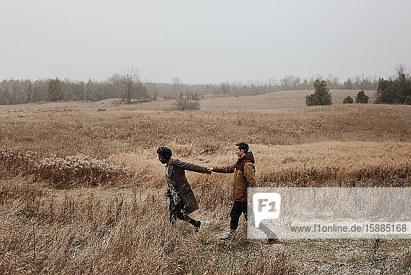Eine schwarze Frau und ein weißer Mann halten sich an den Händen und gehen durch Gräser in einer winterlichen ländlichen Landschaft.