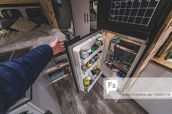 A hand opening the campervan fridge door which is filled with food and jars.