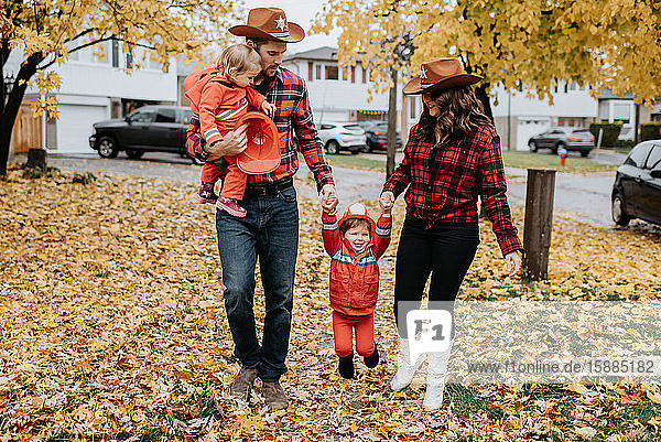 Eine Familie mit zwei Kindern  die für Halloween als Feuerwehrmänner verkleidet sind  spaziert durch einen mit Blättern übersäten Park.