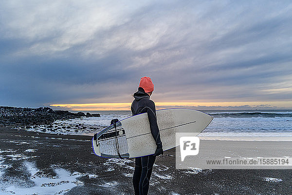 Frau in einem Neoprenanzug am Strand mit einem Surfbrett in der Hand  mit Blick aufs Meer.
