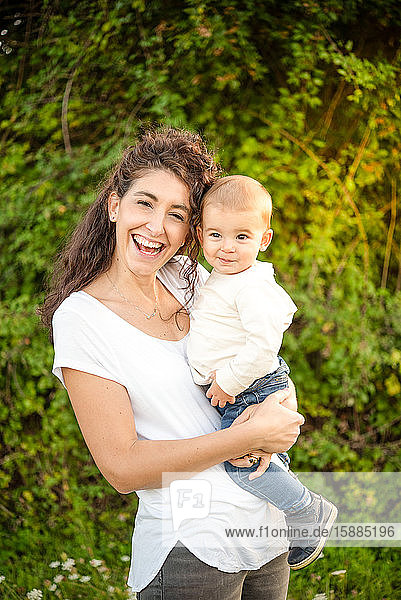 Porträt einer Frau  die einen kleinen Jungen hält  der auf einer Wiese steht und in die Kamera lächelt.