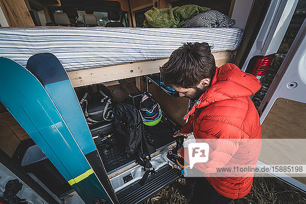 Man standing at the back of a van putting on ski boots standing next to a pair of skiis.