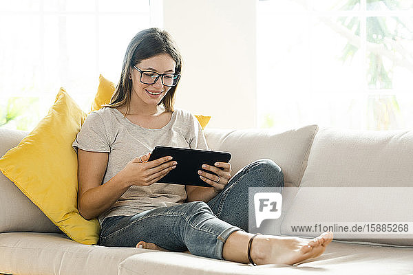 Lächelnde brünette Frau mit Brille  die sich auf der Couch entspannt und auf ihr Tablett schaut