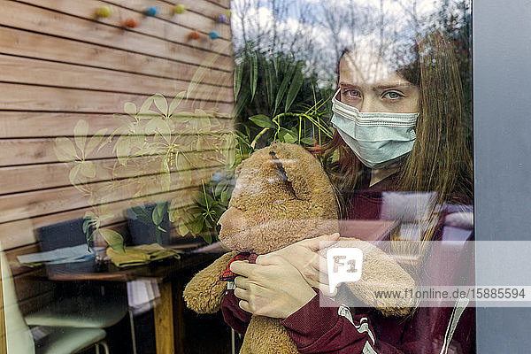 Bildnis eines Mädchens mit chirurgischer Maske und Teddybär hinter Fensterscheibe