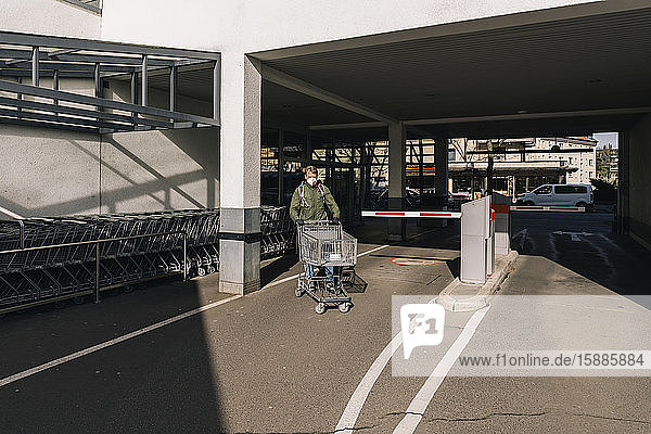 Mann mit Maske schiebt Einkaufswagen vor den Supermarkt