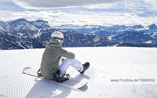 Lächelnde Frau sitzt mit dem Snowboard auf dem Aussichtspunkt und genießt die Aussicht aus der Ferne