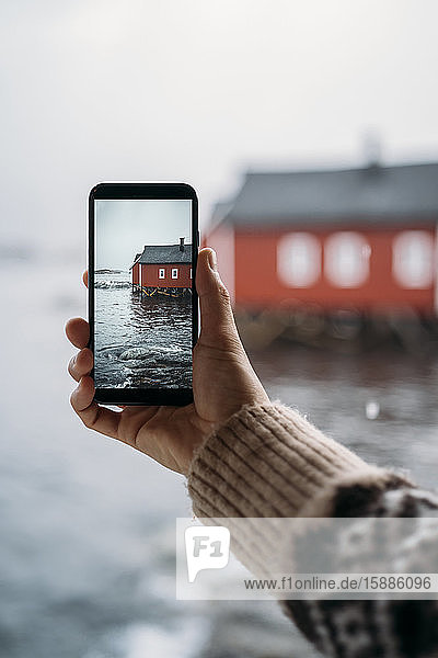 Nahaufnahme eines Touristen  der ein Handyfoto von einer Hütte an der Küste macht  Lofoten  Norwegen