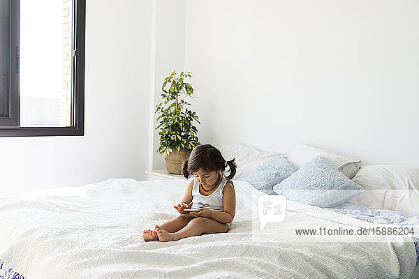 Kleines Mädchen in Unterwäsche sitzt mit Smartphone auf dem Bett