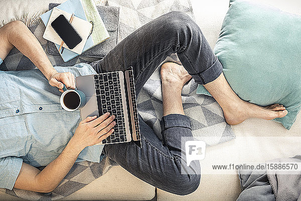Draufsicht auf einen auf dem Sofa liegenden Mann mit Tasse Kaffee und Laptop