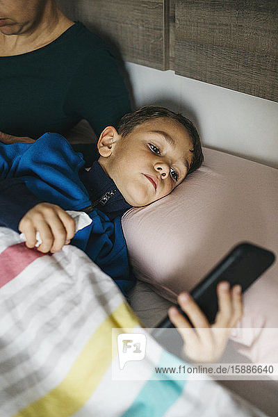 Porträt eines kranken kleinen Jungen  der im Bett liegt und auf ein Smartphone schaut