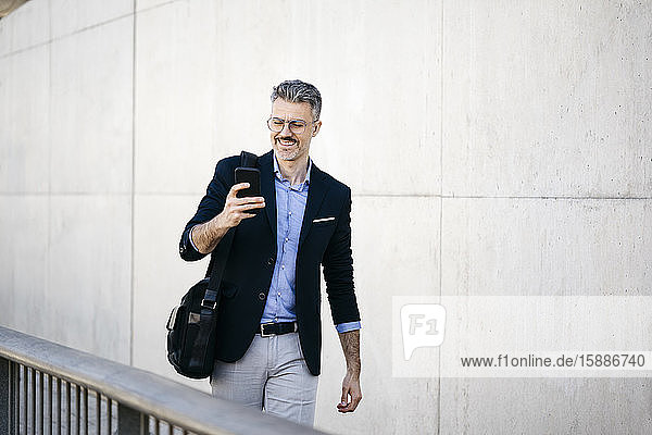 Lächelnder grauhaariger Geschäftsmann geht mit seinem Handy durch die Stadt
