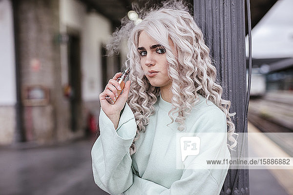 Porträt einer jungen Frau  die auf dem Bahnsteig eine elektronische Zigarette raucht