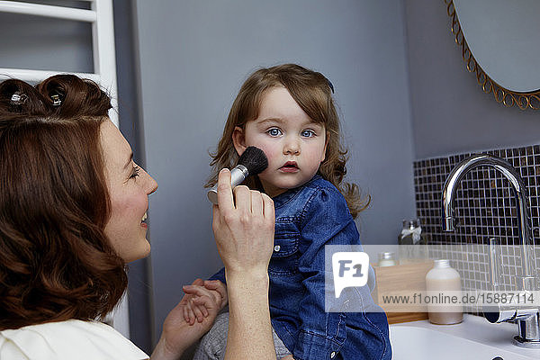 Lächelnde junge Mutter trägt Rouge auf das Gesicht ihrer Tochter im Badezimmer auf