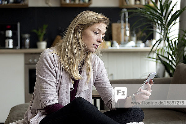 Porträt einer blonden jungen Frau  die zu Hause auf der Couch sitzt und auf ihr Handy schaut