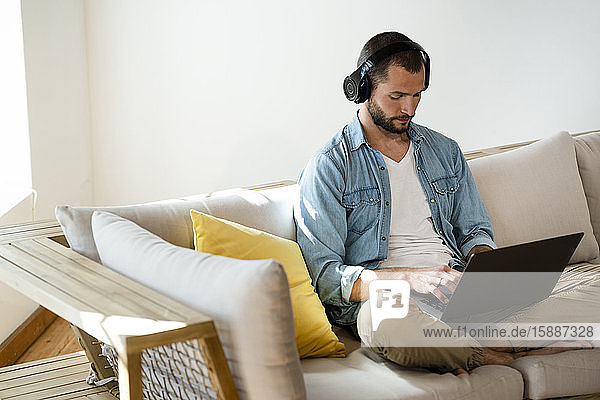 Junger Mann sitzt zu Hause auf dem Sofa mit Kopfhörern und Laptop