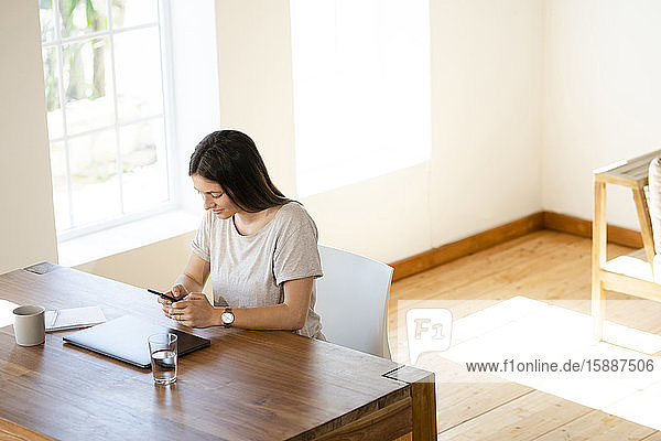 Junge Frau zu Hause sitzt am Tisch und kontrolliert ihr Smartphone im Home-Office