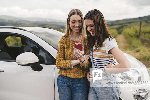 Two happy young women standing beside car sharing cell phone