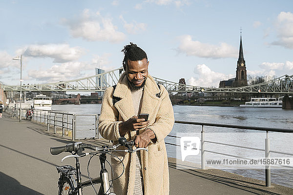 Stylischer Mann mit Fahrrad mit Smartphone am Flussufer  Frankfurt  Deutschland