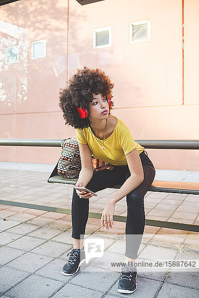 Junge Frau mit Afrofrisur  die an einer Bushaltestelle in der Stadt mit Kopfhörern Musik hört