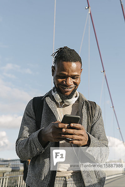 Porträt eines lächelnden Mannes auf einer Brücke mit Kopfhörern und Smartphone