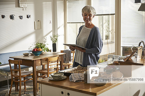Porträt einer lächelnden älteren Frau  die in der Küche mit einem digitalen Tablet steht und Müsli zubereitet