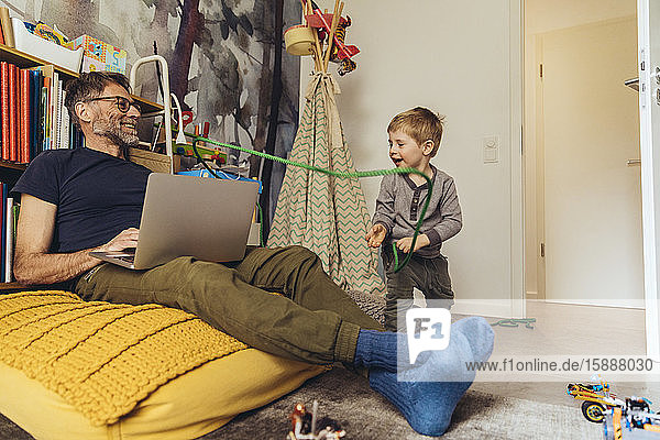 Sohn bindet seinen Vater bei der Arbeit am Laptop im Kinderzimmer
