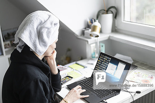 Frau mit Handtuch um den Kopf  die zu Hause am Schreibtisch sitzt und einen Laptop benutzt
