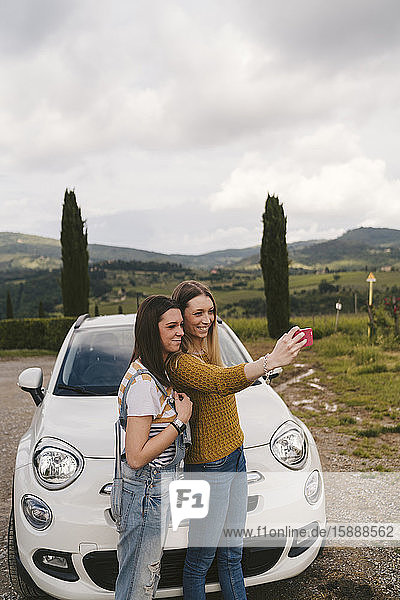 Zwei glückliche junge Frauen  die neben einem Auto stehen und sich ein Selfie nehmen  Toskana  Italien